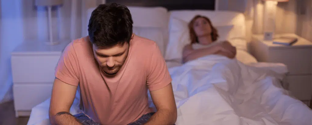 Man in a pink shirt sits on the edge of a bed looking down, while a woman lies asleep under white covers in the background. The room is dimly lit.